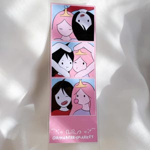 May include: A rectangular bookmark featuring three stacked panels of cartoon illustrations depicting two characters with pink and black hair. The bottom section displays the text @RINABERRYMARKET in white lettering against a solid pink background.