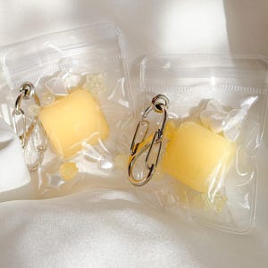 May include: Two clear plastic pouches containing a yellow square-shaped object and small star-shaped beads. Each pouch is attached to a silver-toned metal carabiner clip. The items are arranged side by side, showcasing the yellow square and star accents.