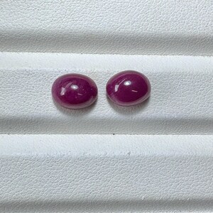 May include: Two oval-shaped, polished gemstones with a deep magenta hue. The gems are displayed on a white, textured surface. The stones have a smooth, reflective surface, suggesting they are cabochons, possibly rubies.