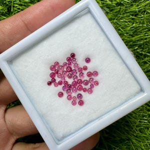 May include: A collection of small, round, pink gemstones displayed in a white square container with a white interior. The gemstones vary in size and are scattered across the white surface. The container is held against a background of green grass.