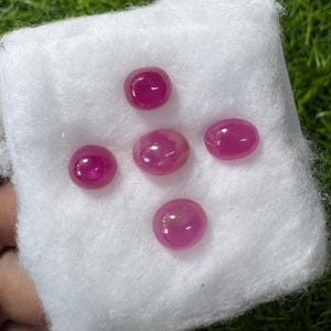May include: Five polished, oval-shaped, ruby gemstones displayed on a white cloth. The rubies are a deep, vibrant red color, with a smooth, reflective surface. The gemstones are arranged in a scattered pattern.