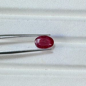 Op de afbeelding: Een dieprode, ovaalvormige robijn, vastgehouden door een zilveren pincet. De robijn wordt getoond tegen een witte, getextureerde achtergrond. Deze afbeelding toont een edelsteen, ideaal voor sieraden.