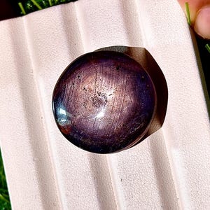 以下が含まれることがあります： 深い紫色の、研磨された丸い宝石。シャトヤンシー効果を示しています。石の表面には、目に見える縞模様と微妙な光沢があります。宝石は、明るい色のテクスチャード加工された背景に置かれています。
