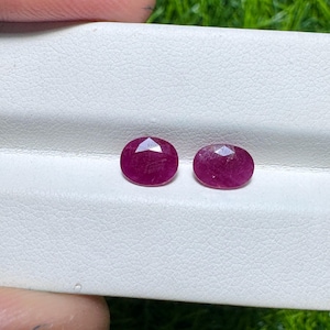 May include: Two oval-cut, faceted ruby gemstones. The rubies are a deep, rich red colour and are displayed on a white textured surface. The gemstones are ready for jewellery making.