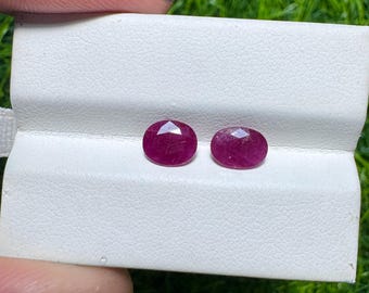 Puede incluir: Dos piedras preciosas de rubí ovaladas y facetadas. Los rubíes son de un color rojo intenso y rico y se muestran sobre una superficie blanca texturizada. Las piedras preciosas están listas para la fabricación de joyas.