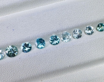 May include: A collection of round, faceted gemstones in varying shades of blue and teal. The gems are arranged in a diagonal line on a white, textured surface, showcasing their brilliance and color variations. The gemstones are of different sizes.