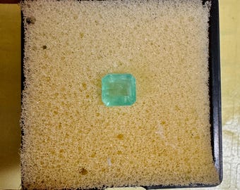 May include: A square-cut, light green emerald gemstone. The gem is set on a yellow foam pad within a black plastic tray. The emerald is a vibrant, clear colour, and the cut is precise. The foam provides a soft background for the gemstone.