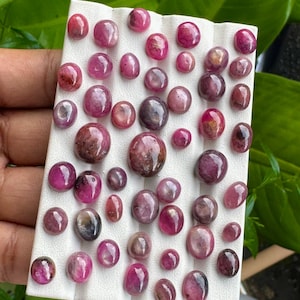 May include: A collection of polished, oval-shaped gemstones in varying shades of pink and red, displayed on a white surface. The gemstones are of different sizes and shapes, with a glossy finish. The background is blurred, with green foliage.