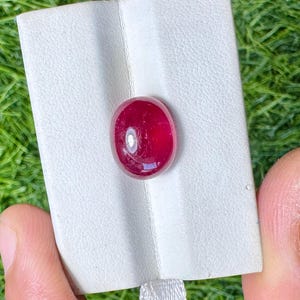 May include: A vibrant, oval-shaped, deep red gemstone. The polished ruby is displayed on a white textured surface, held by a person's fingers. The background is a blurred green.