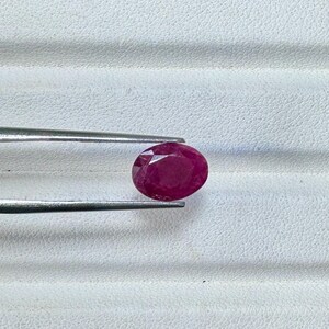 May include: An oval-shaped, deep red gemstone is held by a pair of silver tweezers. The ruby is faceted, reflecting light. The background is a textured, white surface.