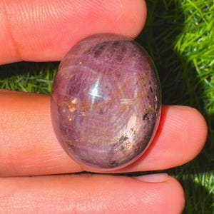 Peut inclure: Une pierre précieuse polie, de forme ovale, d'un violet profond, avec des motifs internes et une surface réfléchissante. La pierre est tenue entre les doigts, mettant en valeur sa beauté naturelle et ses variations de couleur uniques.