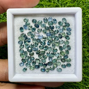 May include: A collection of numerous small, oval-cut loose gemstones in various shades of blue and green, arranged inside a square white display box with a white foam insert. The faceted stones exhibit a mix of teal, navy, and light green hues.
