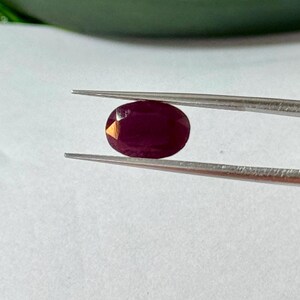 May include: A faceted, oval-shaped ruby gemstone held by tweezers. The ruby is a deep, rich red color, with visible internal inclusions. The tweezers are silver-colored, and the background is a plain white surface.