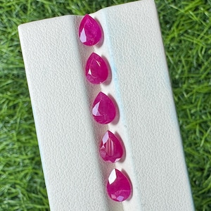 Puede incluir: Cinco rubíes facetados en forma de pera están dispuestos en una línea vertical sobre un soporte de exhibición blanco texturizado. Cada piedra preciosa exhibe un tono rojo rosado profundo y saturado con facetas.
