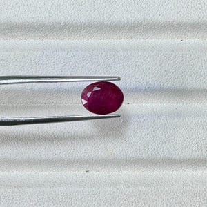 May include: A faceted, oval-shaped ruby gemstone held by silver tweezers. The ruby is a deep, rich red color and is set against a white textured background. The tweezers are positioned to display the gemstone.