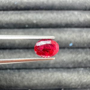 May include: A faceted, oval-shaped ruby gemstone held by tweezers. The ruby is a deep red color with visible internal inclusions. The tweezers are silver-colored, and the background is a dark gray, textured surface.