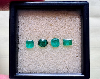 May include: Four vibrant green emerald gemstones of varying shapes are displayed in a black square frame. The gemstones include a square cut, an oval cut, and two other cuts. The gemstones are set against a beige background.
