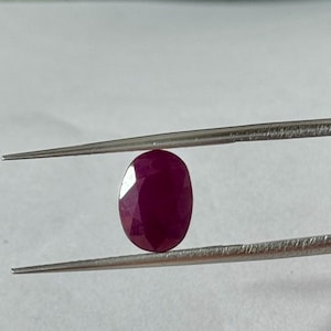 Pode incluir: Uma gema de rubi oval e facetada, segurada por uma pinça. O rubi é de uma cor vermelha profunda e rica, com facetas visíveis que capturam a luz. A pinça é prateada e segura a gema contra um fundo neutro.