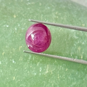 May include: A polished, round, ruby gemstone held by tweezers. The gemstone is a deep, saturated red color with a glossy surface. The background is a light green, textured surface.