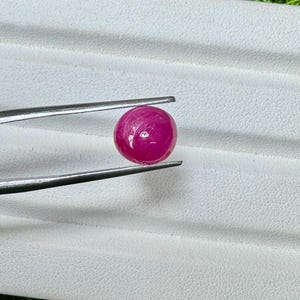 May include: A round, polished, deep pink gemstone is held by a pair of silver tweezers. The gem has a smooth, reflective surface. The background is a white, textured surface with a hint of green.