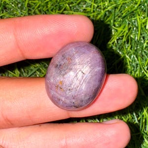 May include: A smooth, oval gemstone in shades of purple and lavender, held in a hand. The stone has a polished surface with subtle internal lines and flecks of darker colour. The background is a vibrant green, suggesting grass or foliage.