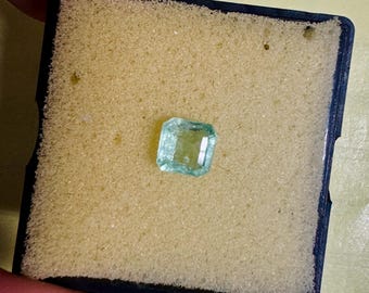 May include: A light green emerald gemstone, rectangular in shape, resting on yellow foam within a dark blue frame. The gem is clear and faceted, revealing its internal structure. The image is a close-up, emphasizing the emerald's clarity and colour.