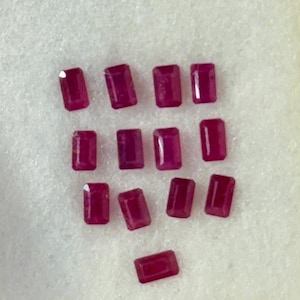 May include: A collection of rectangular, deep red gemstones, possibly rubies, arranged on a white, textured surface. The gems have a polished appearance and are of varying sizes. The overall presentation suggests a collection of precious stones.