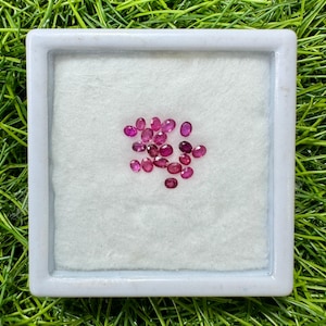 May include: A collection of oval-shaped, deep red gemstones displayed in a white square container with a soft, white interior. The gemstones are scattered across the surface, showcasing their polished facets. The container sits on a bed of green grass.
