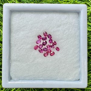 Op de afbeelding: Een verzameling ovaal geslepen, dieproze edelstenen gepresenteerd in een witte tray. De edelstenen liggen op een wit, getextureerd oppervlak, in een witte lijst, tegen een groene achtergrond.