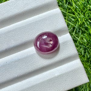 Op de afbeelding: Een gepolijste robijn edelsteen met cabochon-slijpsel. De edelsteen heeft een diepe, rijke rode kleur met een glad, afgerond oppervlak. De robijn ligt op een wit, getextureerd oppervlak, met groen gras op de achtergrond.