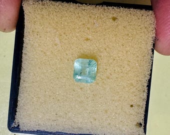 May include: A square-cut, light blue-green gemstone is presented in a black case with a yellow foam insert. The gemstone is the focal point, showcasing its clarity and colour. The case is held by a person.