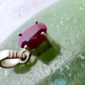 May include: A pendant featuring a deep red, oval-cut gemstone secured in a four-prong silver setting. The piece includes a polished silver bail with vertical grooved detailing, designed for attachment to a chain or cord for wear as jewelry.