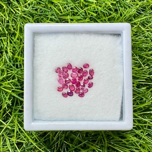 May include: A collection of oval-cut, vibrant red gemstones displayed in a white square container. The gemstones are arranged on a white, textured surface, set against a backdrop of green grass.