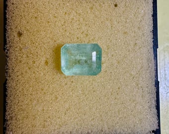 May include: A rectangular, light green emerald gemstone. The gem is set against a yellow foam background within a black square frame. The emerald has a clear, reflective surface.