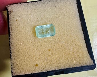 May include: A rectangular, pale blue gemstone rests on a yellow foam pad inside a black frame. The gem is translucent, with internal features visible. The image highlights the gem's clarity and colour, presented in a professional setting.
