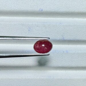 May include: A polished, oval-shaped, red gemstone is held by a pair of silver tweezers. The gemstone has a smooth surface and a deep, rich color. The tweezers are positioned against a white, textured background.
