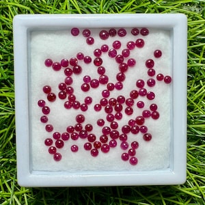 May include: A collection of small, round, deep red gemstones, possibly rubies, are displayed inside a white square container with a white interior. The container sits on a bed of green grass, creating a contrasting background.