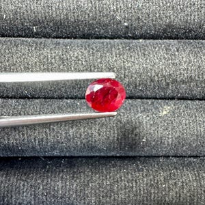 May include: A faceted, oval-shaped, deep red ruby gemstone is held by silver tweezers against a dark gray background. The ruby's surface reflects light, showcasing its brilliance. This gemstone is likely for jewelry making.