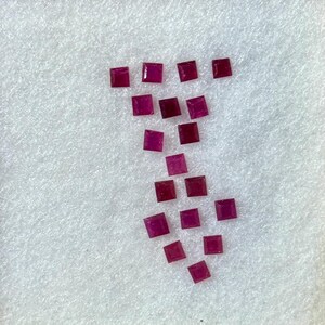 Peut inclure: Une collection de pierres précieuses carrées, rouge foncé, possiblement des rubis, disposées sur une surface blanche et texturée dans un cadre de couleur claire. Les pierres sont de taille et de forme uniformes.