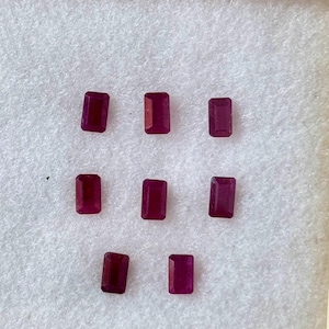 May include: Eight rectangular, deep red gemstones are arranged on a white surface within a wooden frame. The gems have a polished, reflective surface, and are evenly spaced. The gems are likely rubies, and are suitable for jewelry making.