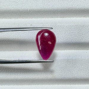 May include: A teardrop-shaped, polished ruby gemstone, held by silver tweezers. The ruby is a deep, saturated red with a smooth, reflective surface. The gemstone is set against a white, textured background.