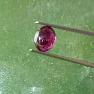 May include: A round, polished, deep red gemstone held by tweezers against a green background. The gem has a reflective surface and appears to be a ruby. The tweezers are silver-colored and hold the gem securely.