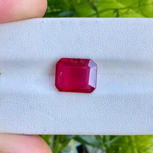 May include: A radiant, rectangular-cut ruby gemstone, presented on a white textured surface. The ruby displays a deep red colour and is set against a backdrop of green foliage. The gemstone is the focal point of the image.