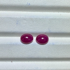 May include: Two oval-shaped, polished ruby gemstones. The rubies are a deep, rich red color with a smooth, glossy surface. They are set against a white, textured background.
