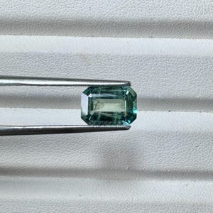 Puede incluir: Una gema rectangular con talla esmeralda y degradado de color verde y turquesa. La gema está sujeta por unas pinzas plateadas sobre un fondo blanco texturizado. Las facetas de la gema reflejan la luz, mostrando su corte.