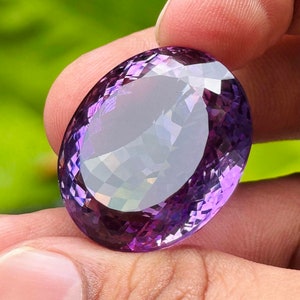 May include: A large, faceted oval amethyst gemstone. The gem is held between fingers, showcasing its deep purple and lavender hues. The cut of the stone creates multiple reflective surfaces, enhancing its brilliance.