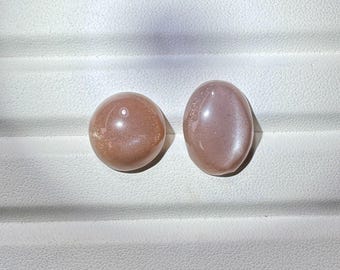 May include: Two polished, oval-shaped gemstones with a shimmering, iridescent surface. One is round, the other is oval. The stones have a warm, peachy-pink color and are set against a white background.
