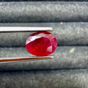 May include: A faceted, oval-shaped ruby gemstone held by tweezers. The ruby is a deep, saturated red color, with visible internal inclusions. The tweezers are silver-colored, and the background is a dark, textured surface.