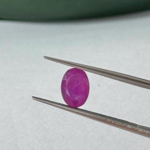 Pode incluir: Uma gema de rubi em forma oval, segurada por uma pinça. O rubi é de cor rosa profunda e a pinça é prateada. A gema é exibida sobre um fundo branco, realçando seu brilho natural.