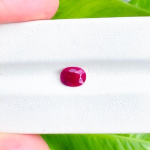 Puede incluir: Un rubí de forma ovalada, presentado sobre una superficie blanca y texturizada. El rubí exhibe un rico color rojo intenso, contrastando con el fondo blanco. La gema es el elemento central, destacando sus facetas pulidas y su brillo.